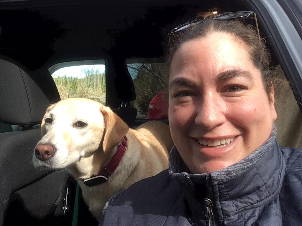 Selfie photo of Jen with her yellow lab, Lyco, over her right shoulder in a vehicle.